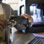 Close up male designer examining robotic hand prototype at laptop next to 3D printer