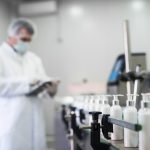 Close up of liquid soap going on a production line in front of cosmetics fabric worker typing on a tablet.